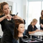 A cosmetology student focusing on cutting hair on a mannequin head during hands-on training at a modern beauty school salon, with an instructor supervising in the background.