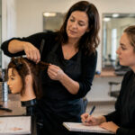 Experienced beauty instructor guiding a student through mannequin hair training in a cosmetology classroom.