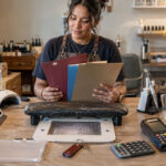 Independent nail professional reviewing three folders for insurance, taxes, and business registration at an actively used rented salon station with financial tools and nail equipment.