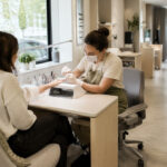 Professional nail technician working with a client at an ergonomic manicure station featuring elevated hand support, organized tools, accessible storage drawers, task lighting, and source-capture ventilation.