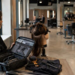Beginning cosmetology student opens a professional training kit beside a mannequin station while observing an advanced student performing supervised hair services on a client.