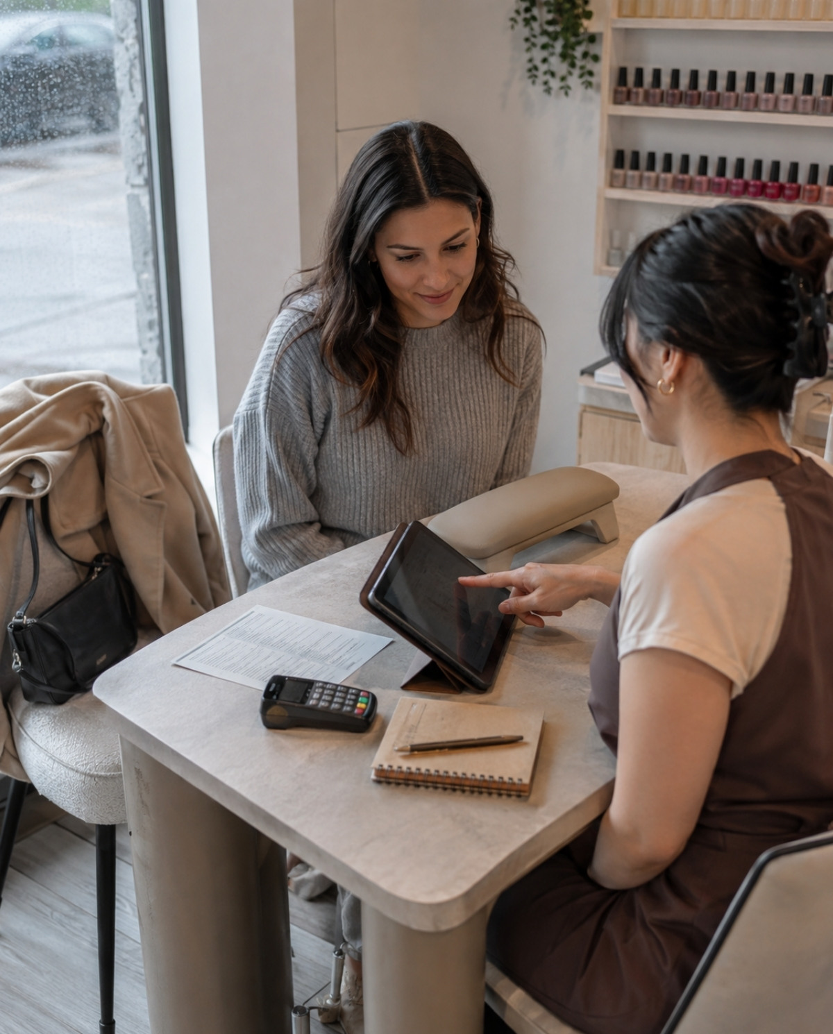 Nail technician discusses appointment expectations with a client using a booking tablet beside a salon policy sheet, payment terminal, and appointment notebook.