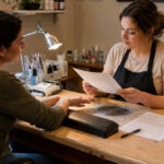 Nail technician reviews a client consultation form while inspecting natural nails at a professional manicure station with a dust collector, curing lamp, e file, hand rest, and organized tools.