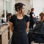 Prospective cosmetology student standing between a mannequin practice station and an active client service area while considering professional beauty training.