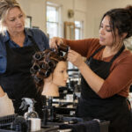 Instructor trainee demonstrates roller placement and hair tension on a mannequin as two adult learners watch and an experienced educator supervises.